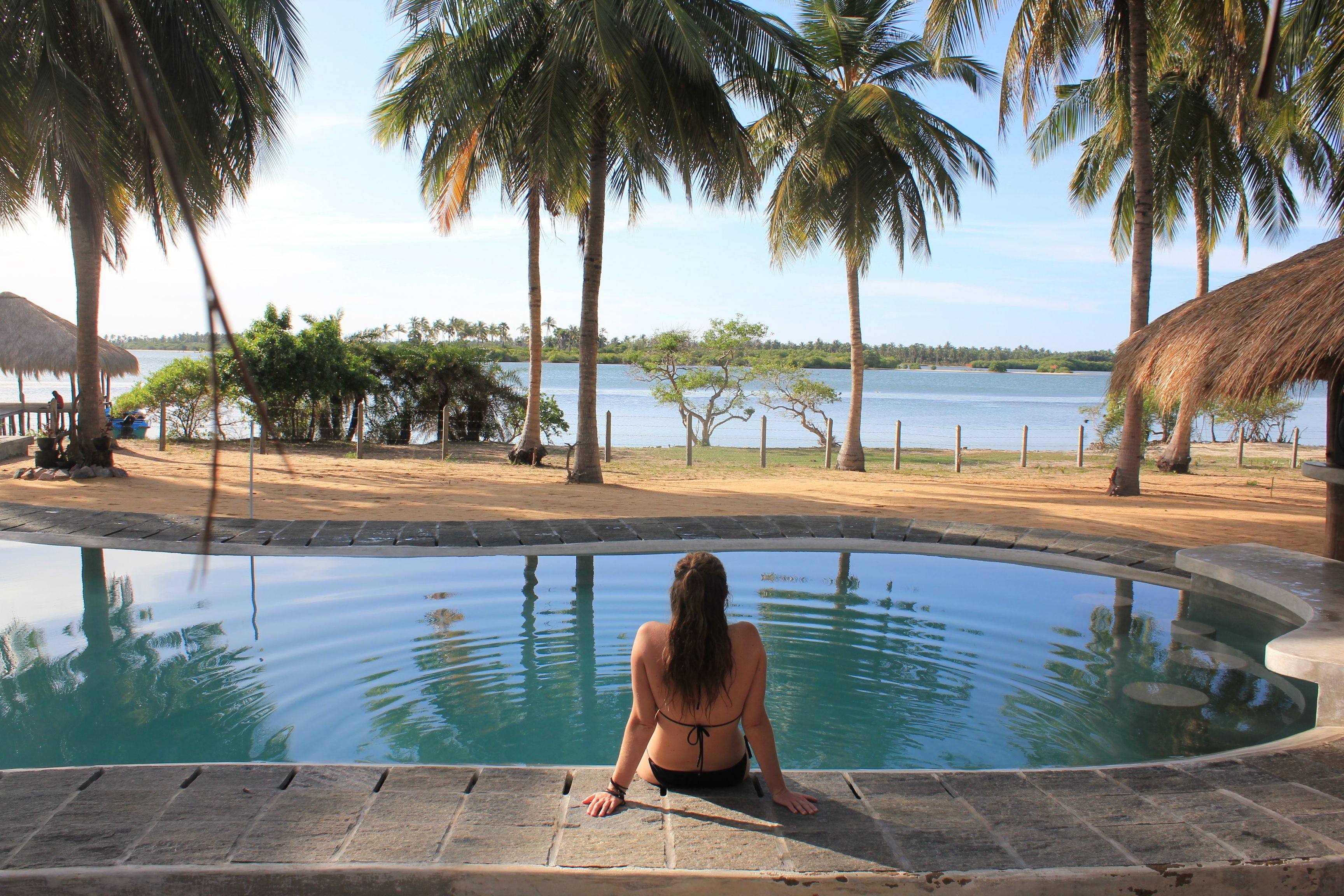 A woman sitting by a pool with a view of the water and palm trees.