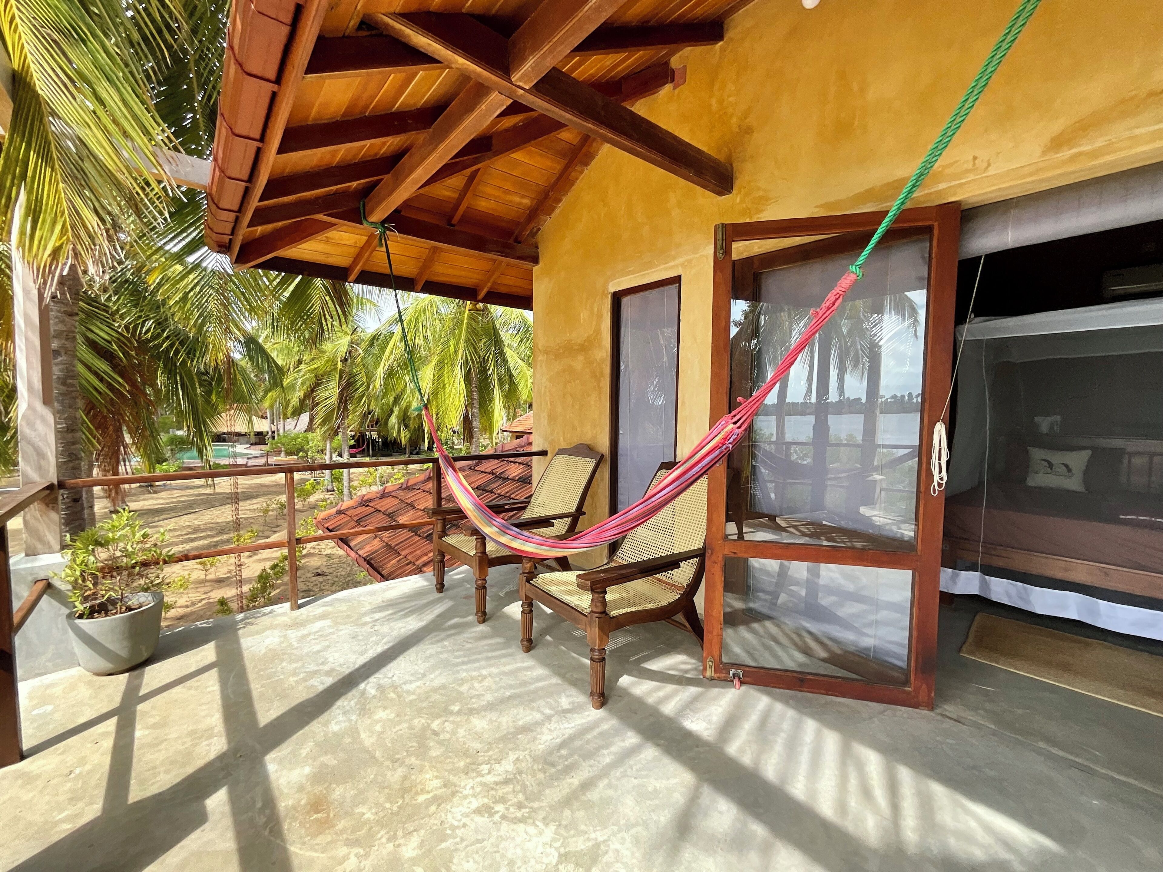 Un espacio al aire libre techado con una hamaca, sillas y vistas a palmeras y una piscina.