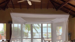 Una habitación con cama bajo un dosel blanco, ventanas amplias con vista al mar y un ventilador de techo.