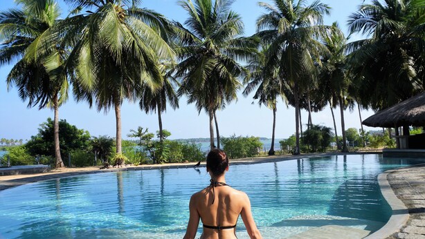 Una mujer con traje de baño negro sentada al borde de una piscina cristalina, con palmeras al fondo.