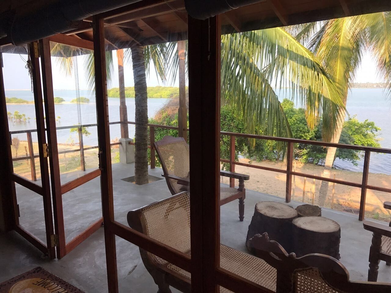 A wooden-framed balcony with wicker furniture overlooking a beach and palm trees.