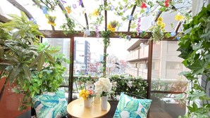 A balcony with a wooden table and two chairs, surrounded by lush green plants and hanging flowers. - Orange House (Osaka)