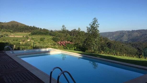 Piscina con muelle de madera y barandillas de metal, rodeada de vegetación exuberante y vistas a colinas distantes.