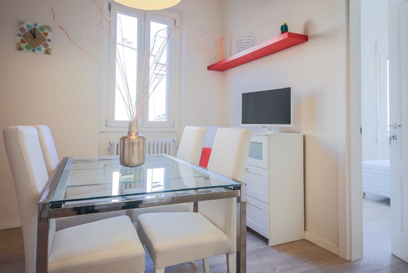 A modern dining area with a glass table, white chairs, a vase, a red shelf, and a television.