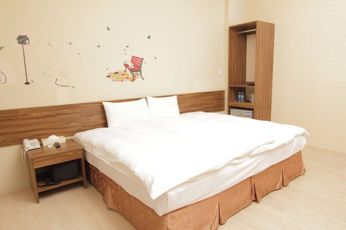 Una cama bien hecha con sábanas blancas, una mesita de noche de madera con un teléfono y un control remoto, y un armario de madera con un espejo.