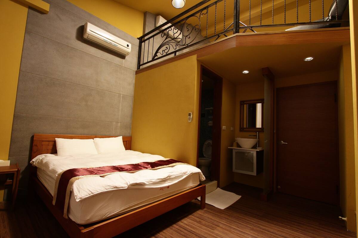 Habitación de hotel con cama de madera, balcón con barandilla de hierro forjado, baño con lavamanos y espejo, y equipo de aire acondicionado en la pared.