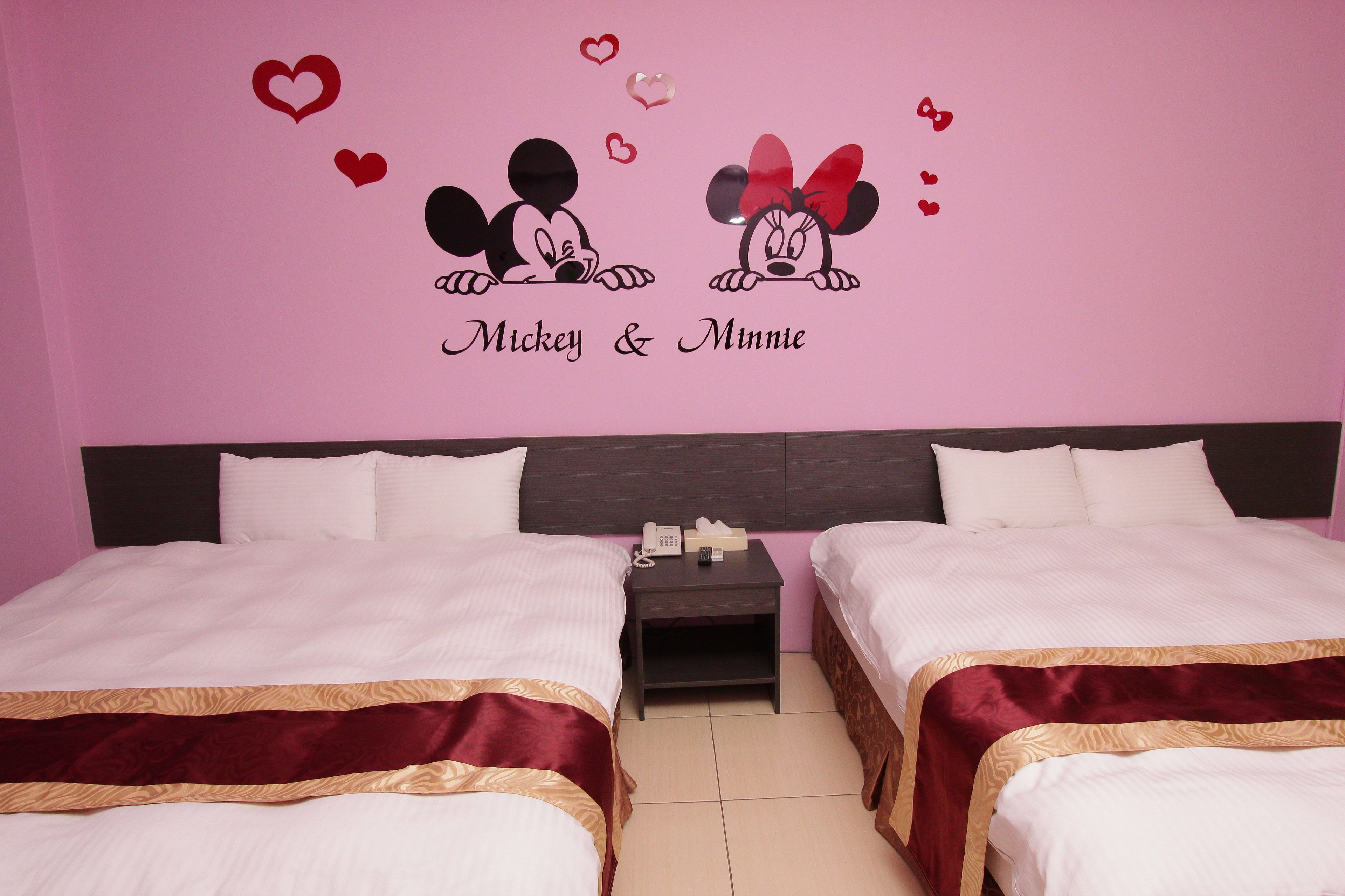 A hotel room with two beds, a nightstand with a phone, and a wall decoration featuring Mickey and Minnie Mouse surrounded by hearts.