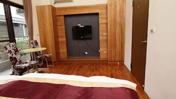 A hotel room with a bed, a TV mounted on a wooden panel, a small table with chairs, and a balcony with a view.