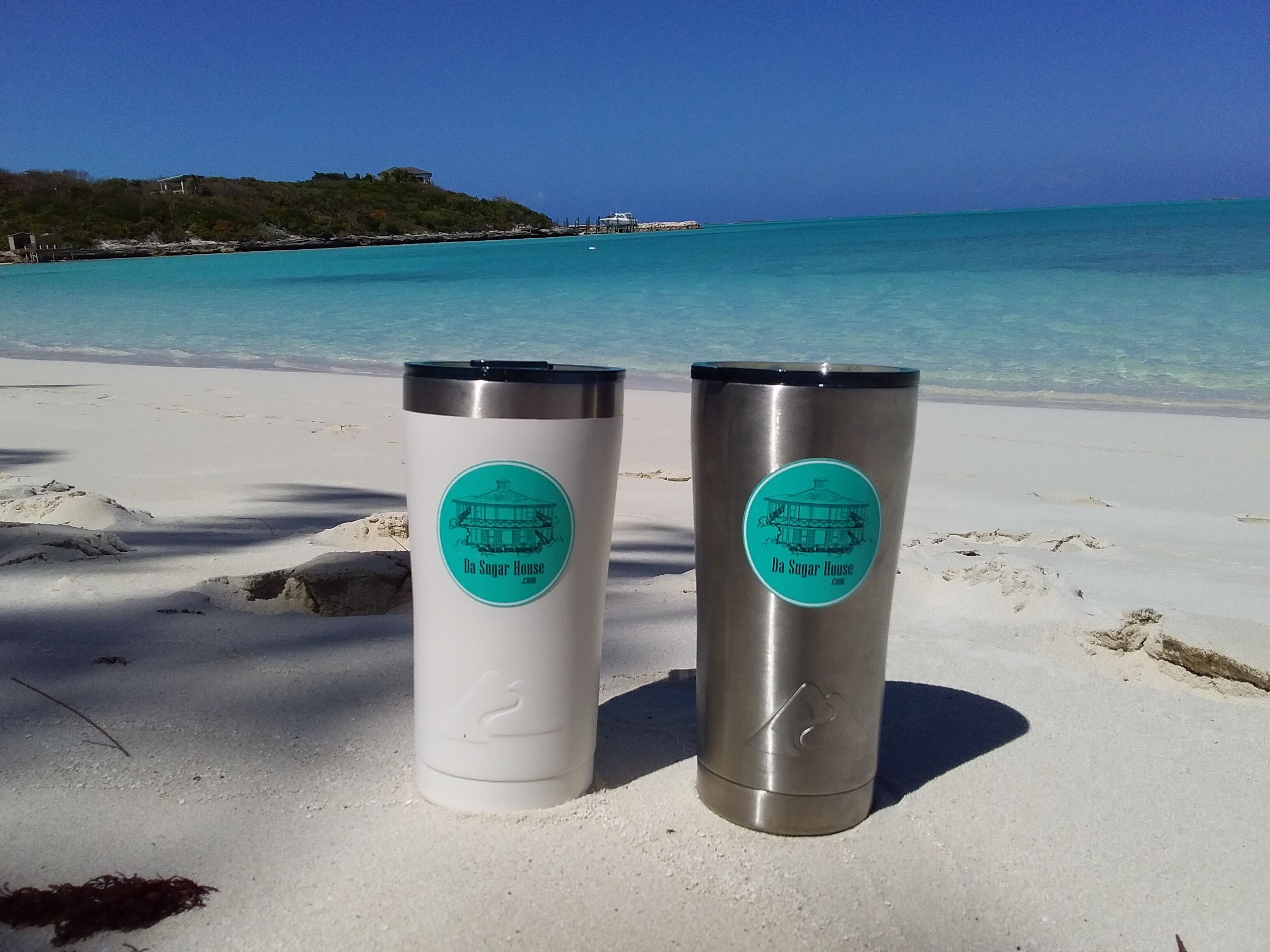 Two stainless steel tumblers with 'Da Sugar House' branding on a sandy beach with clear turquoise water and a distant shoreline.