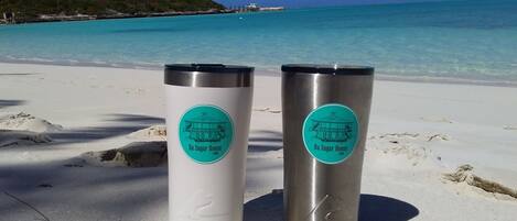 Two stainless steel tumblers with 'Da Sugar House' branding on a sandy beach with clear turquoise water and a distant shoreline.
