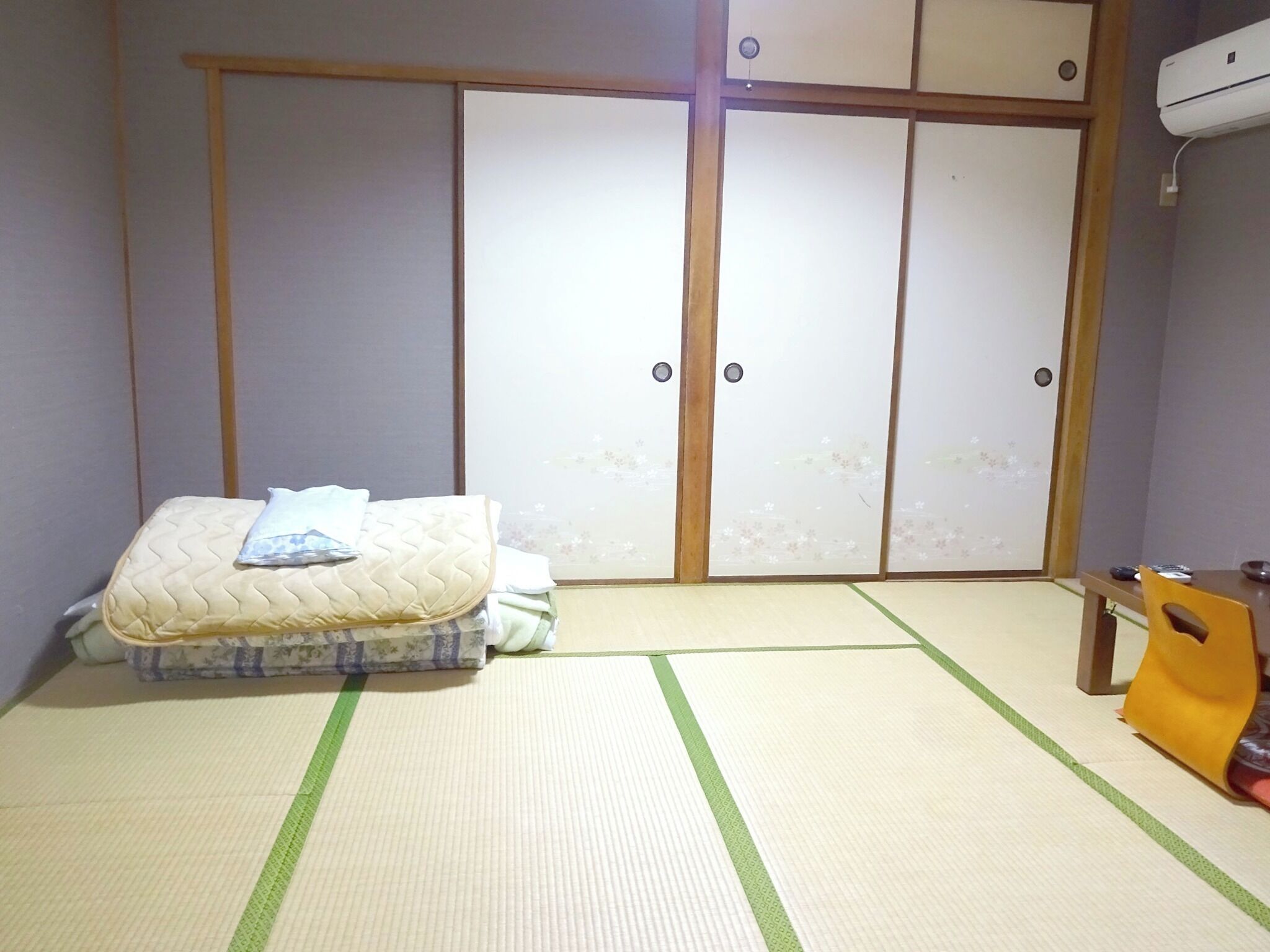 A room with tatami flooring, a futon bed, a wardrobe with sliding doors, and a small table with a chair.