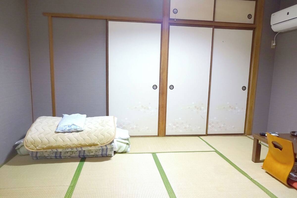 A room with tatami flooring, a futon bed, a wardrobe with sliding doors, and a small table with a chair.