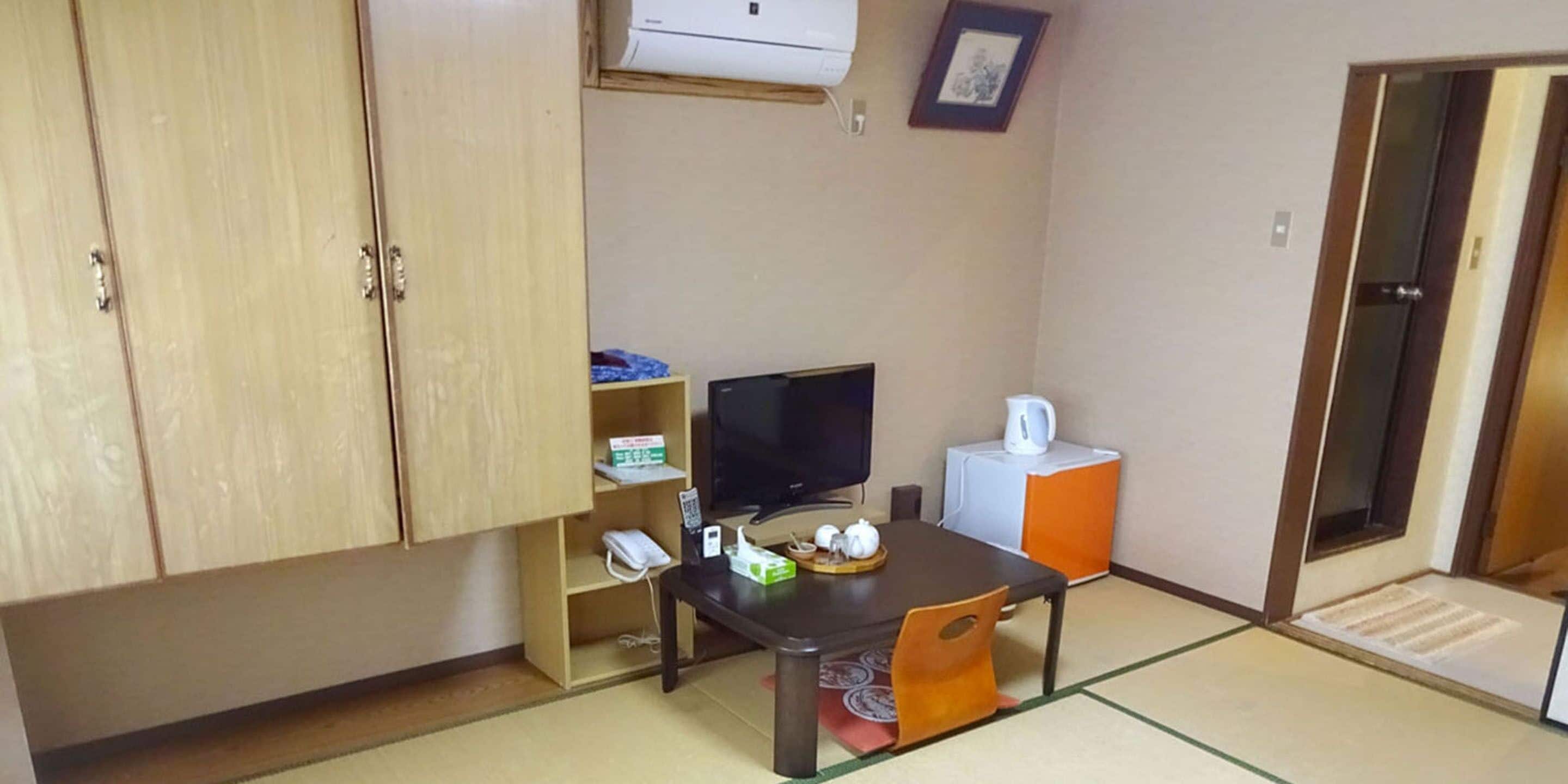 Une pièce de taille modeste, avec une armoire en bois, une petite télévision, une table avec une chaise, une bouilloire blanche et un tableau encadré accroché au mur.