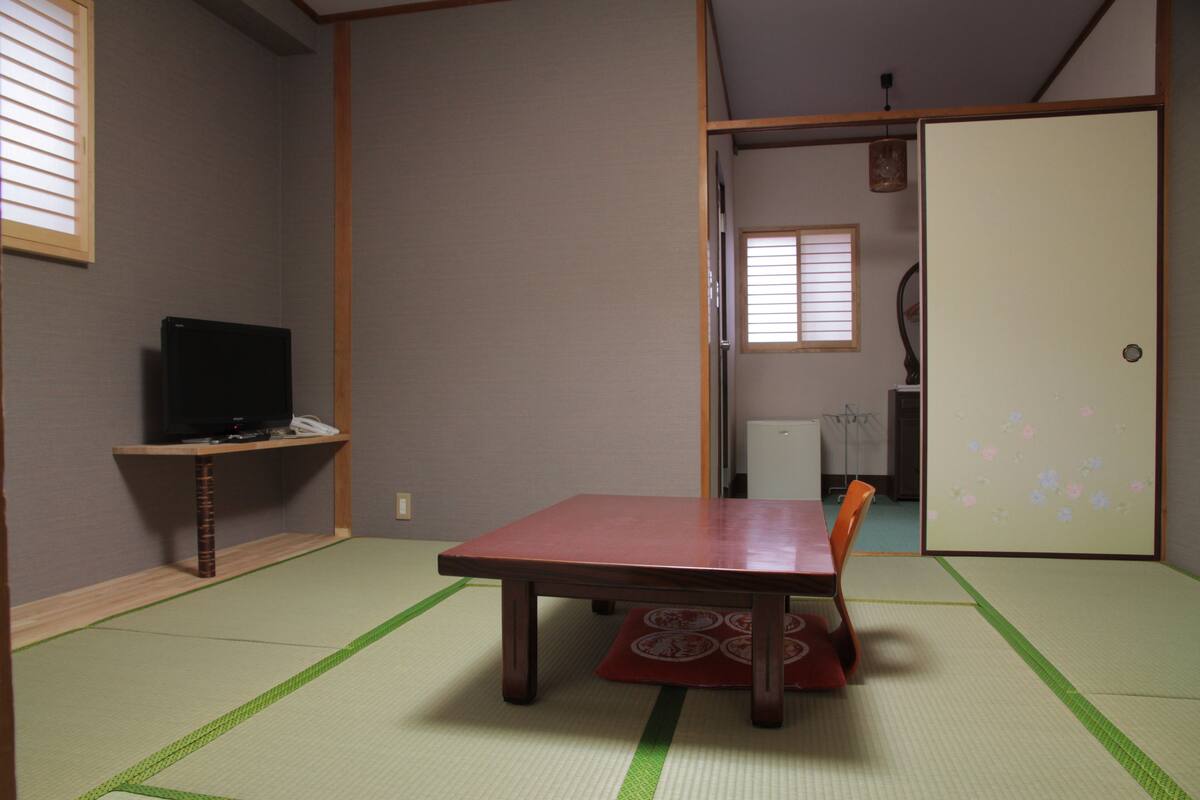 A traditional Japanese room with tatami flooring, a low table, a television on a shelf, and sliding doors.