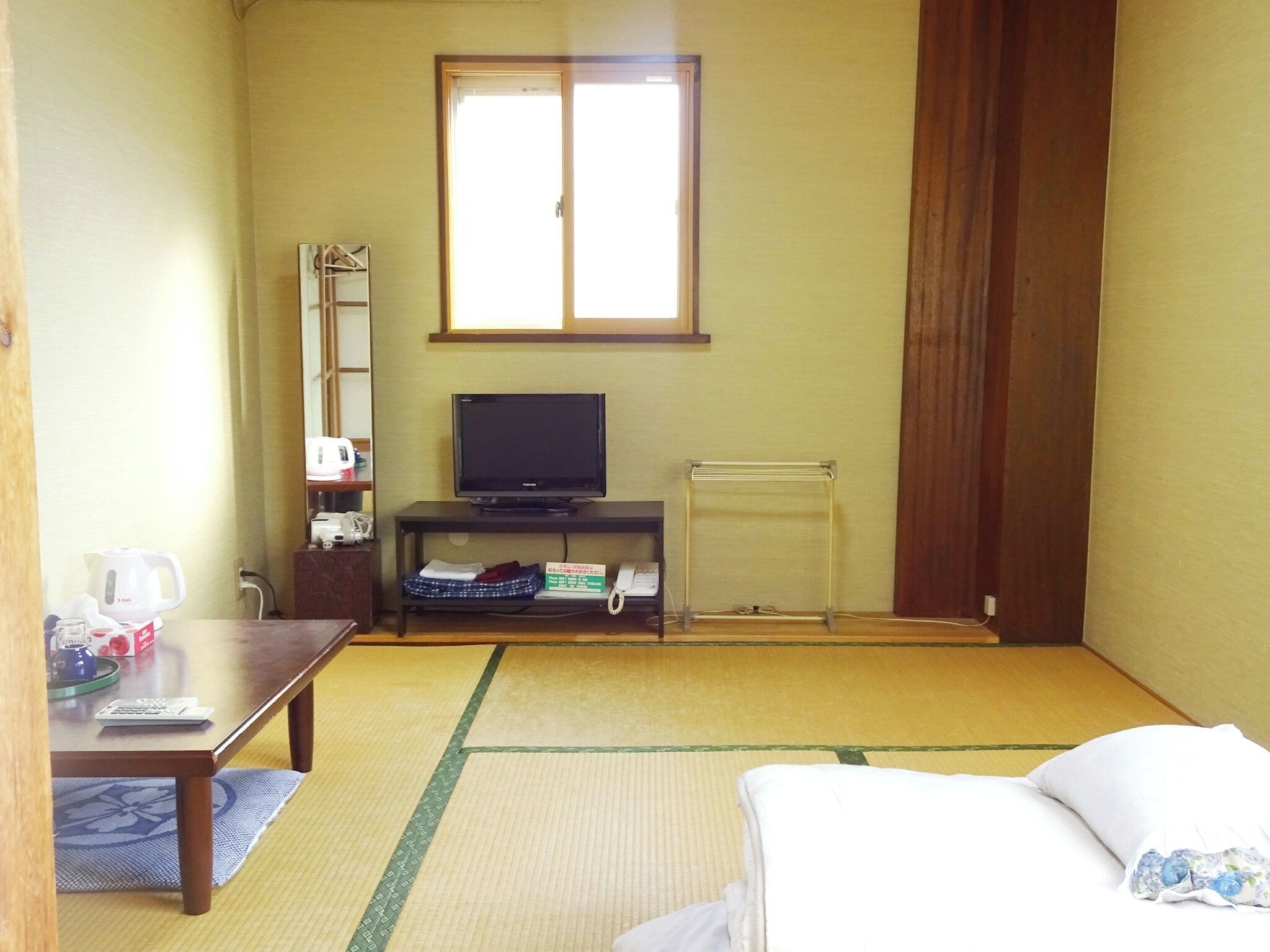A room with tatami flooring, a bed, a TV on a stand, a kettle, a telephone, and a window with curtains.