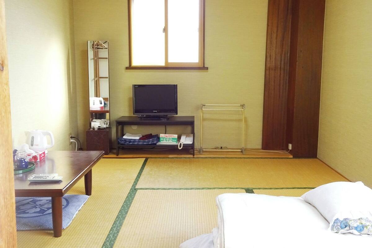 A room with tatami flooring, a bed, a TV on a stand, a kettle, a telephone, and a window with curtains.