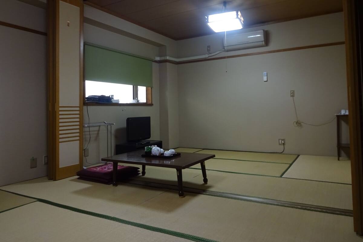 A traditional tatami mat room with a tatami mat floor, a low table with a cushion, a television, and a ceiling-mounted air conditioner.