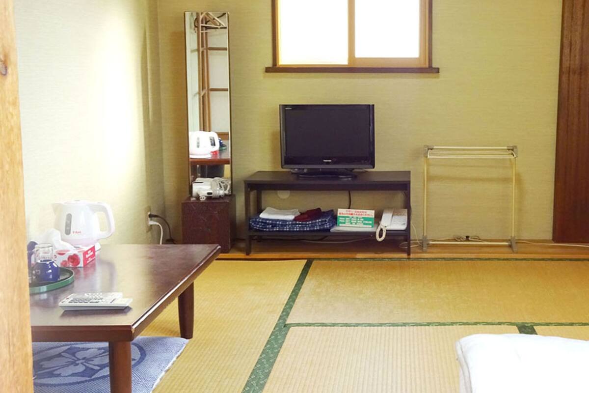 A room with tatami flooring, a bed, a TV on a stand, a small table with a kettle and cups, and a shelf with books and a box.