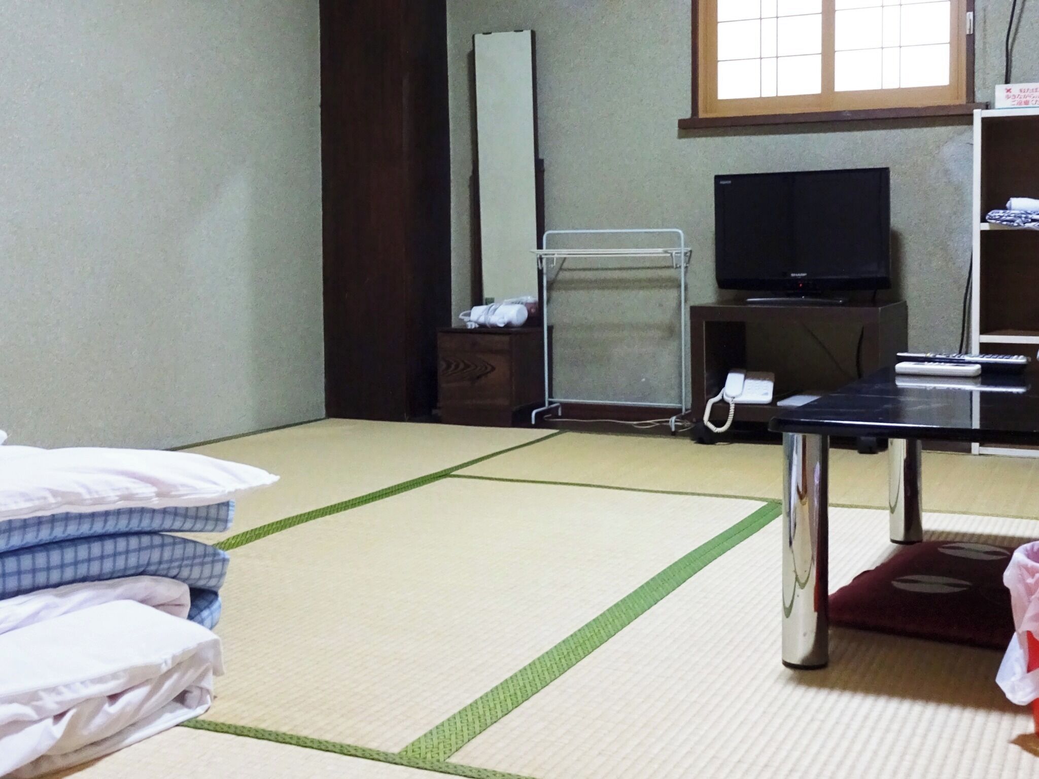 A room with tatami flooring, a bed with pillows, a desk with a telephone, a television, and a window with a view.