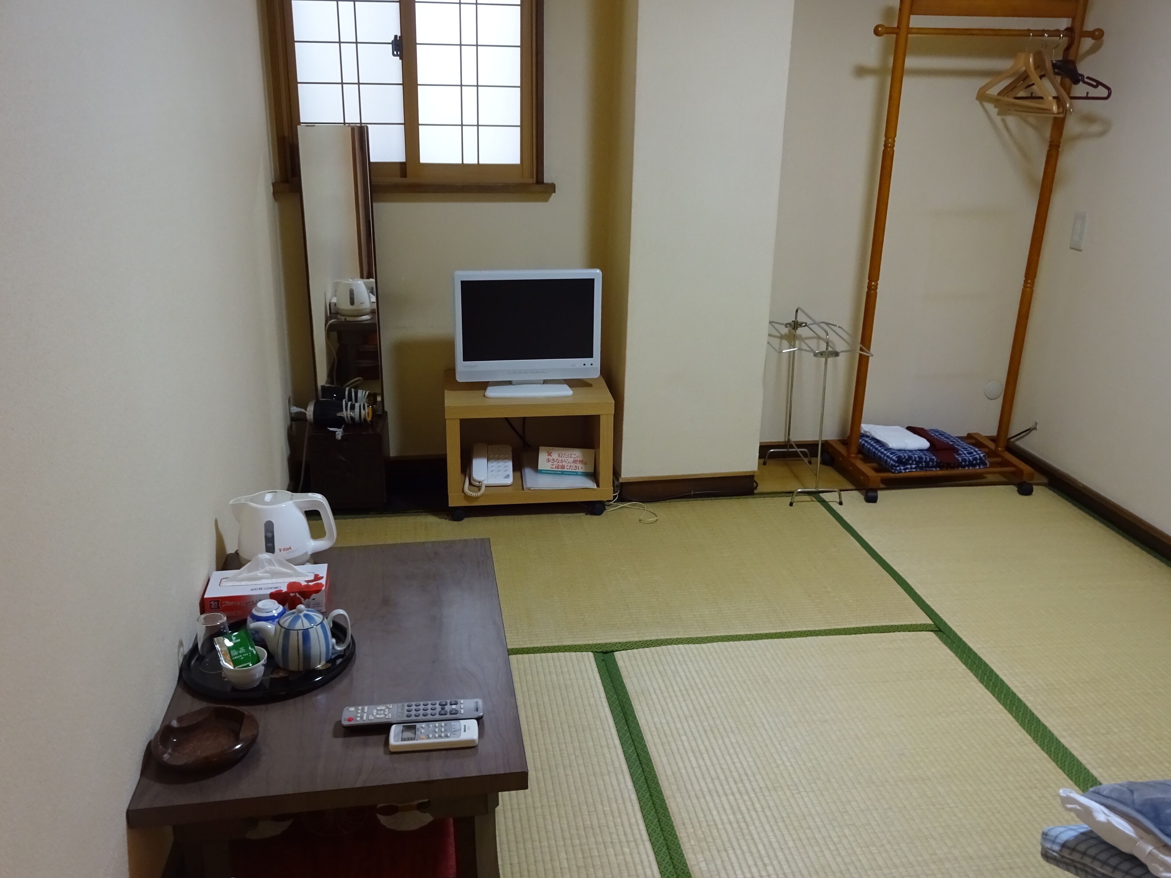 Ein Zimmer mit Tatami-Boden, einem Fernsehgerät auf einem Standfuß, einem Holztisch mit Fernbedienungen, einem Wasserkocher und einer Tasse.