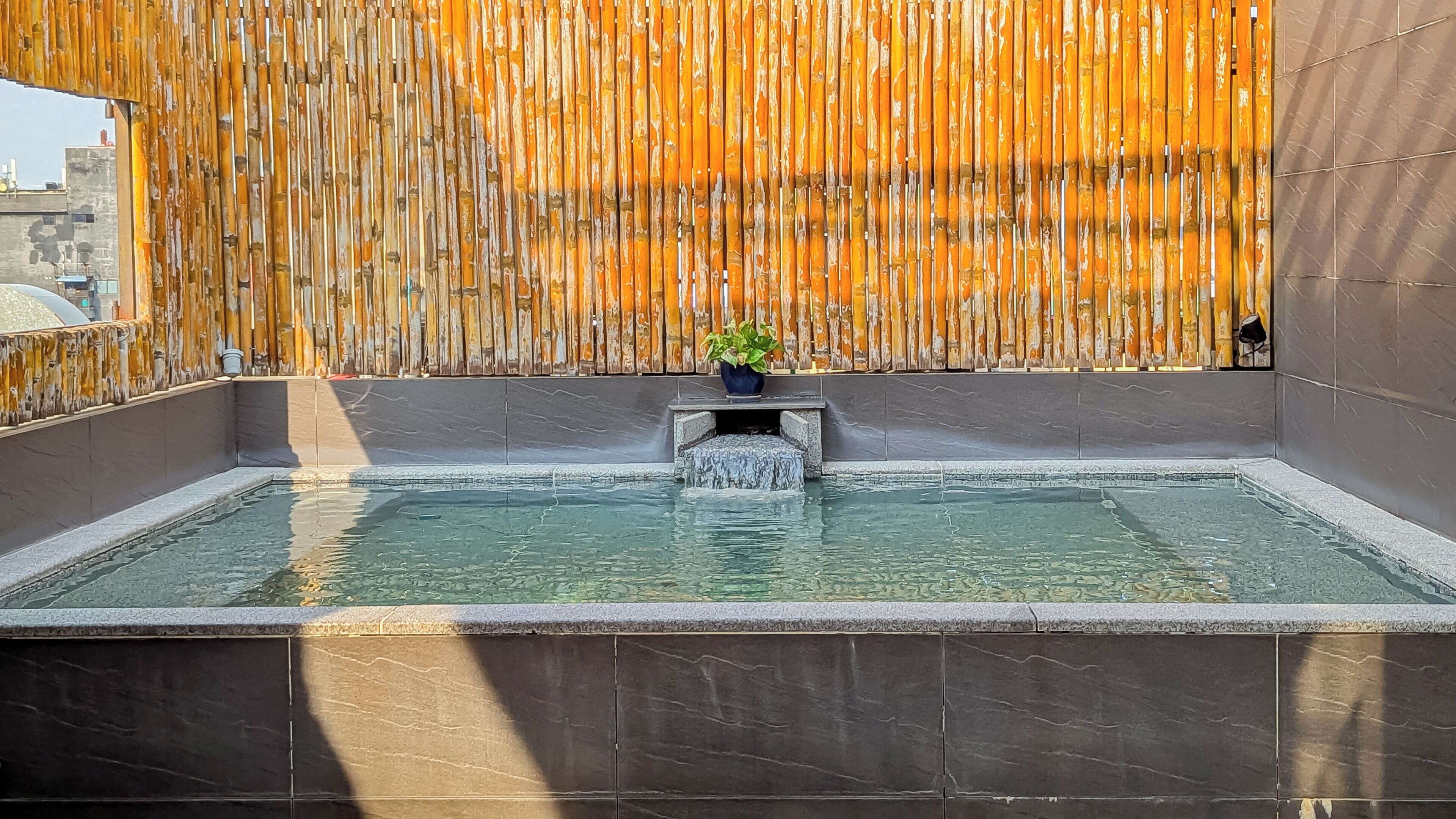 Une piscine rectangulaire, avec un mur en bambou en arrière-plan, une petite cascade et une plante en pot au centre.