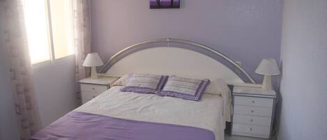 A bedroom with a bed, two bedside tables, a nightstand, a window with curtains, and a wall-mounted painting.