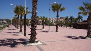 A paved pedestrian walkway lined with palm trees and street lamps, leading towards a building complex in the background. - Enjoy and relax with the light, the murmur of the sea and the fantastic views (San Juan de los Terreros)