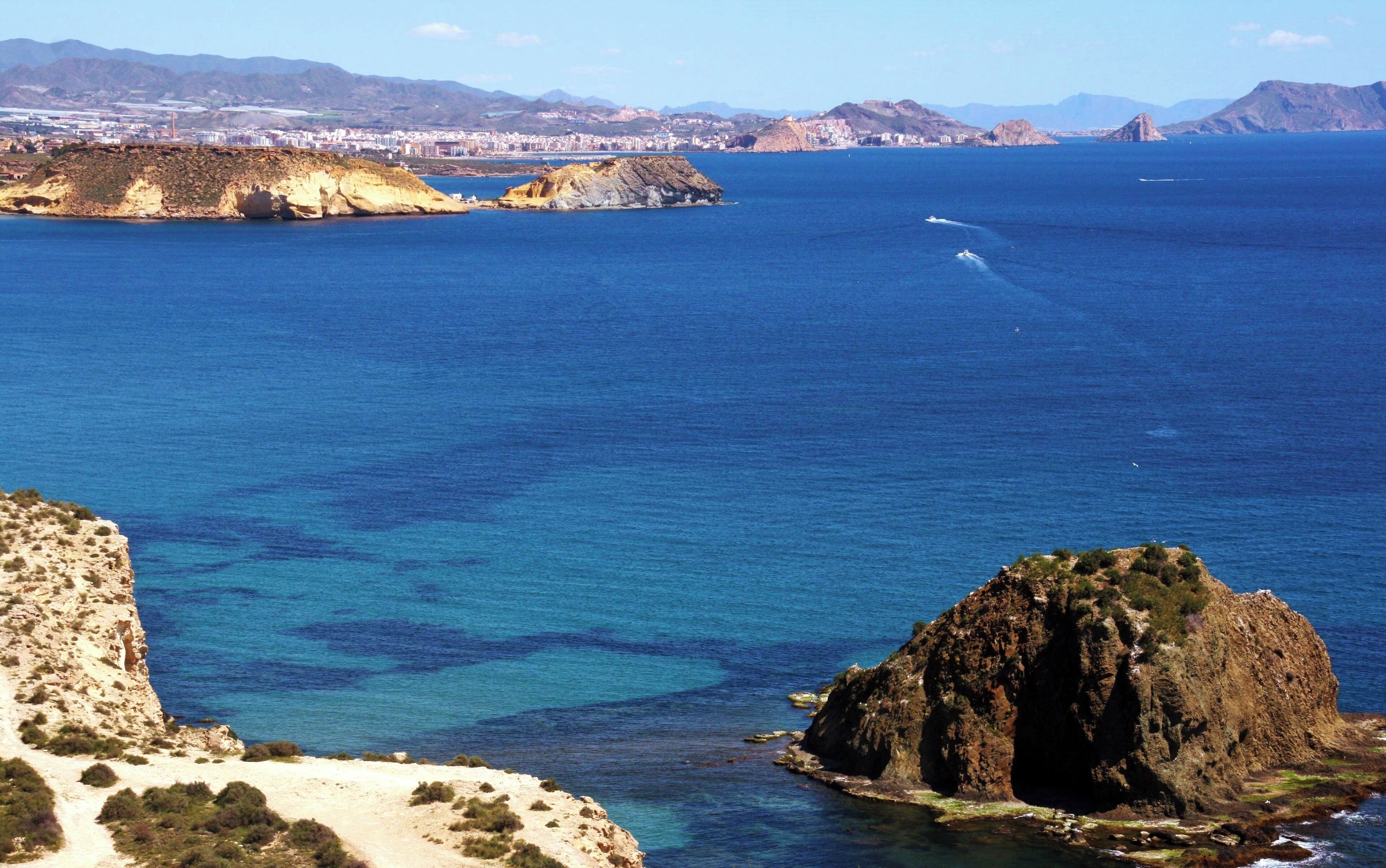 A coastal landscape with clear blue waters, rocky formations, and a distant cityscape.