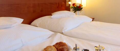 Una cama bien hecha con ropa blanca, una bandeja con productos de desayuno como croissants, frutas y una cafetera, y un florero con flores sobre la mesita de noche.