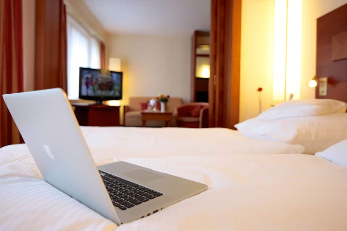 A hotel room with a bed, a laptop on the bed, a TV in the background, and a small table with a lamp.