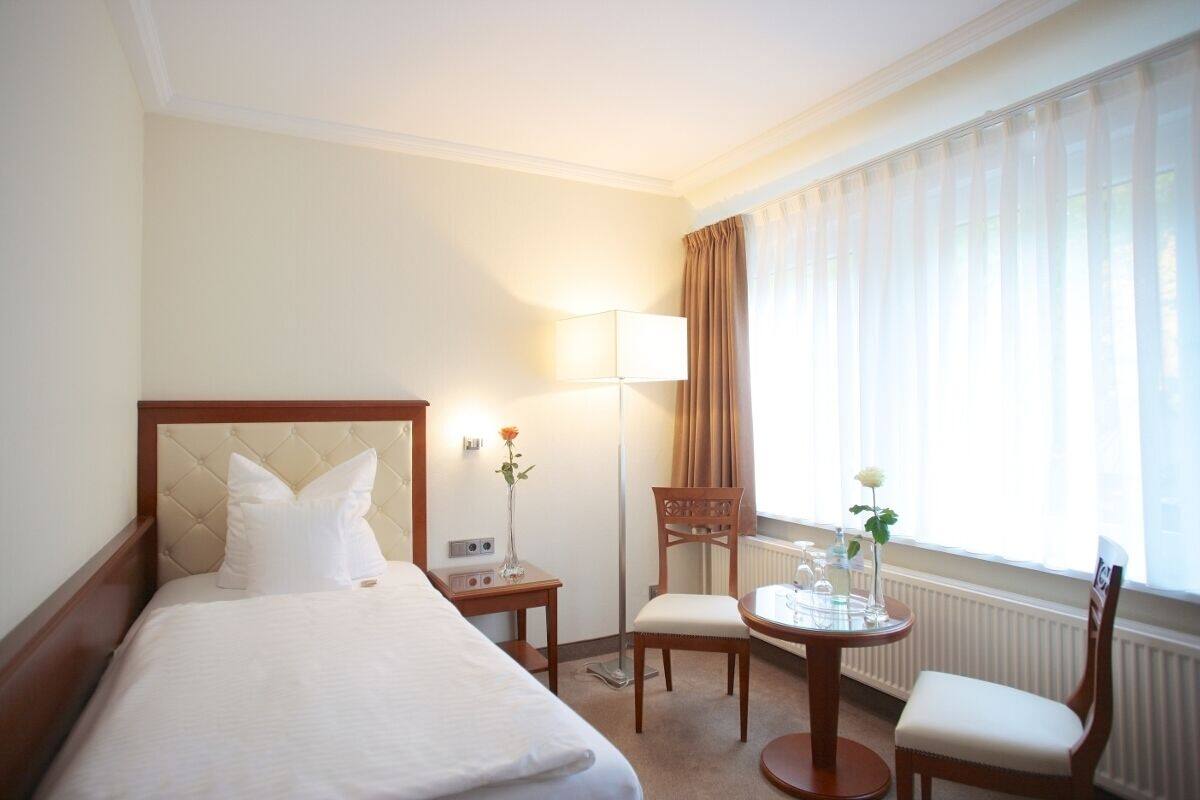A hotel room with a bed, a bedside table with a lamp, a chair, and a small table with a vase of flowers.
