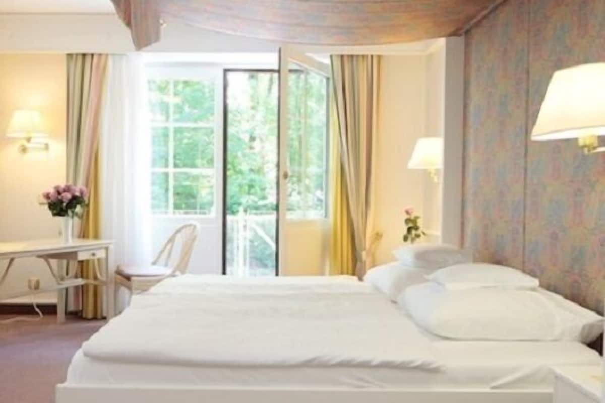 A hotel room with a large bed, a desk, and a chair. There is a window with curtains, and a view of greenery outside.