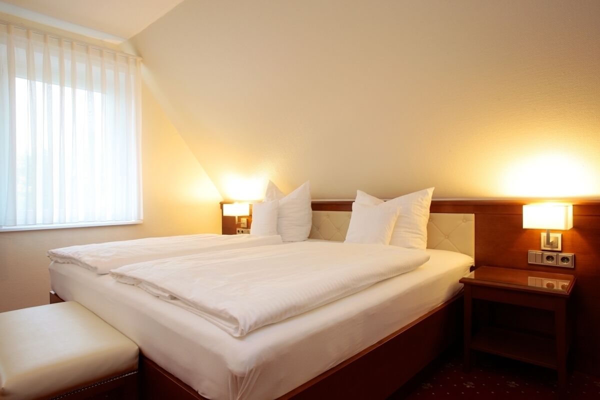 Habitación de hotel con una cama grande, dos mesitas de noche con lámparas, un banco y una ventana con persianas.