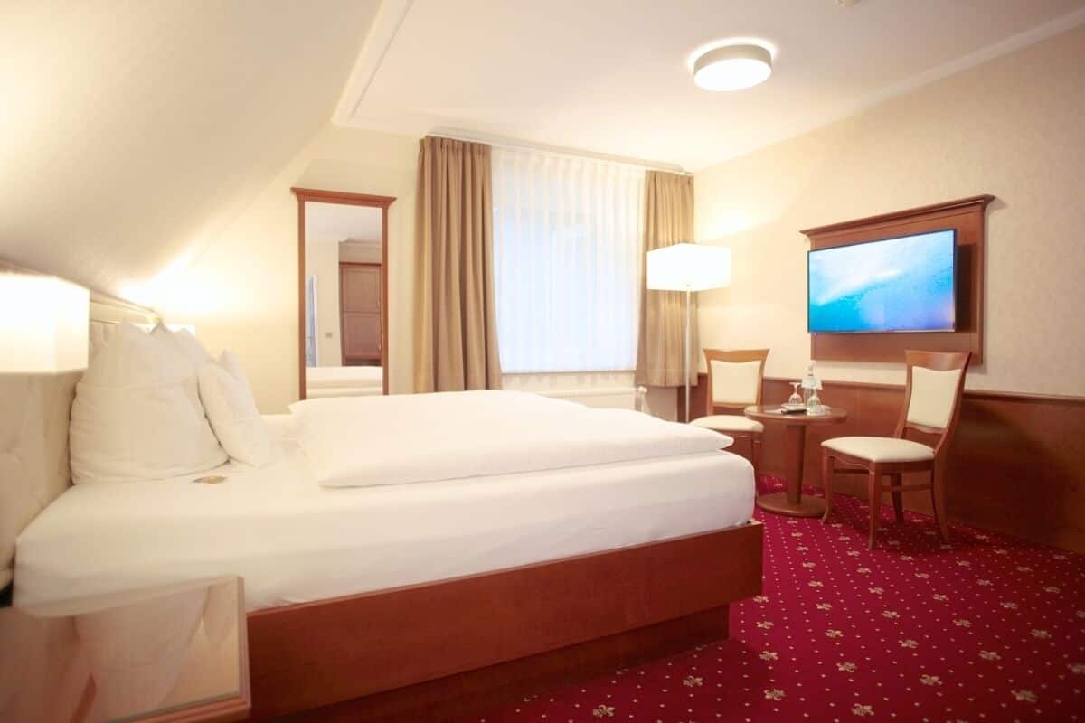 A hotel room with a large bed, two bedside tables, a TV mounted on the wall, and a red carpet.