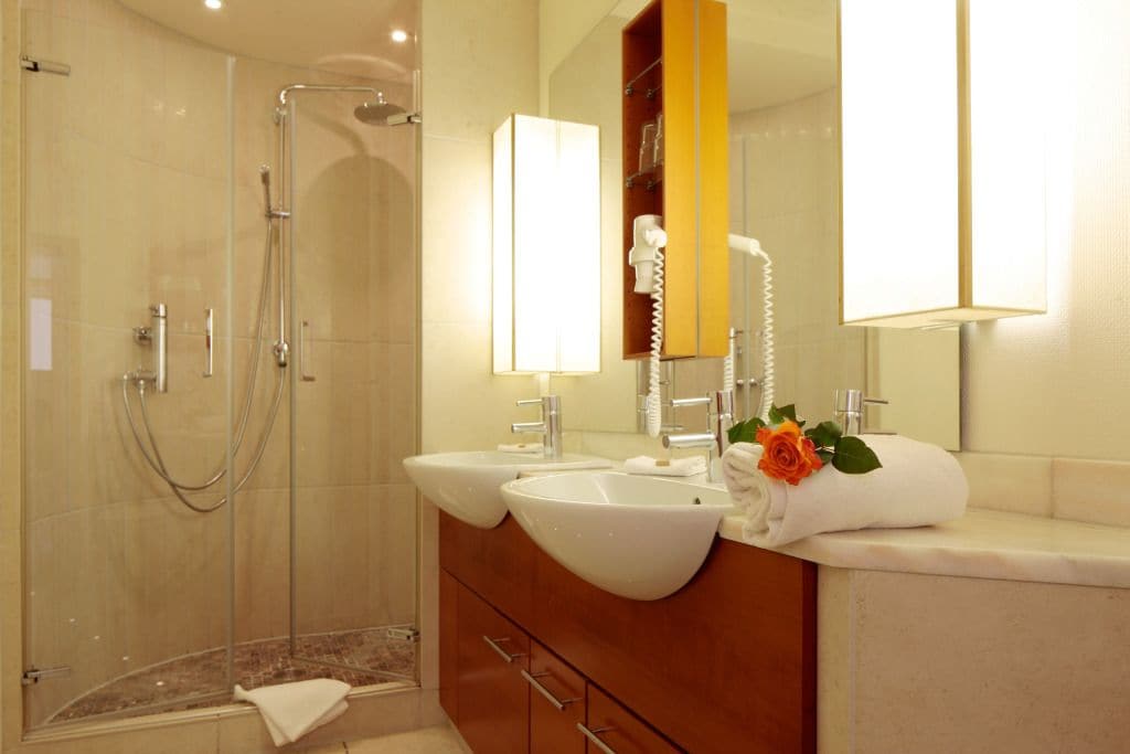 A modern bathroom with a glass-enclosed shower, two sinks, a vanity with a mirror, and a towel with a rose on the counter.