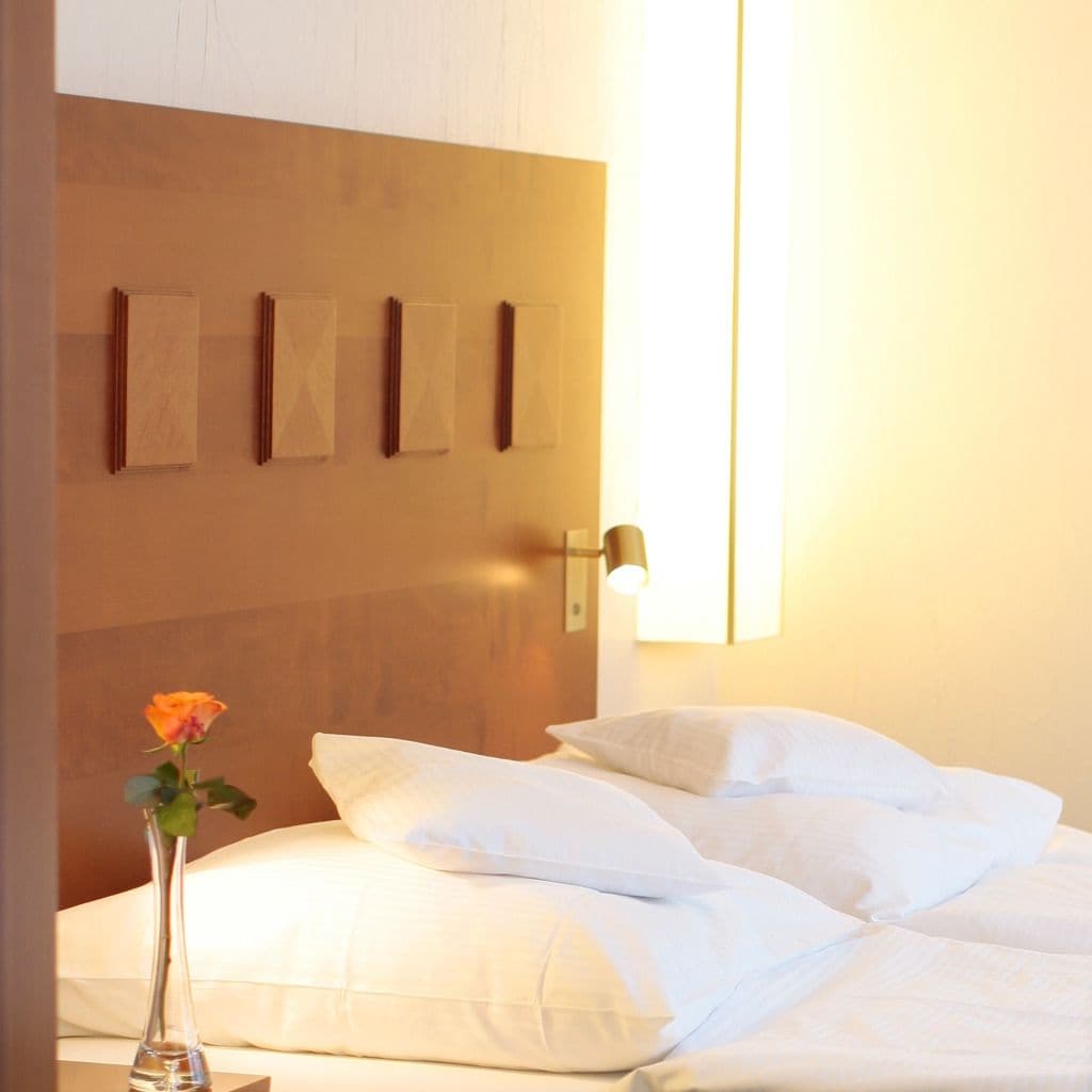 A hotel room with a neatly made bed, a vase with a single flower, and a wall-mounted light fixture.