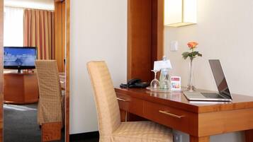 A hotel room with a wooden desk, a laptop, a telephone, a vase with a single flower, and a television in the background.