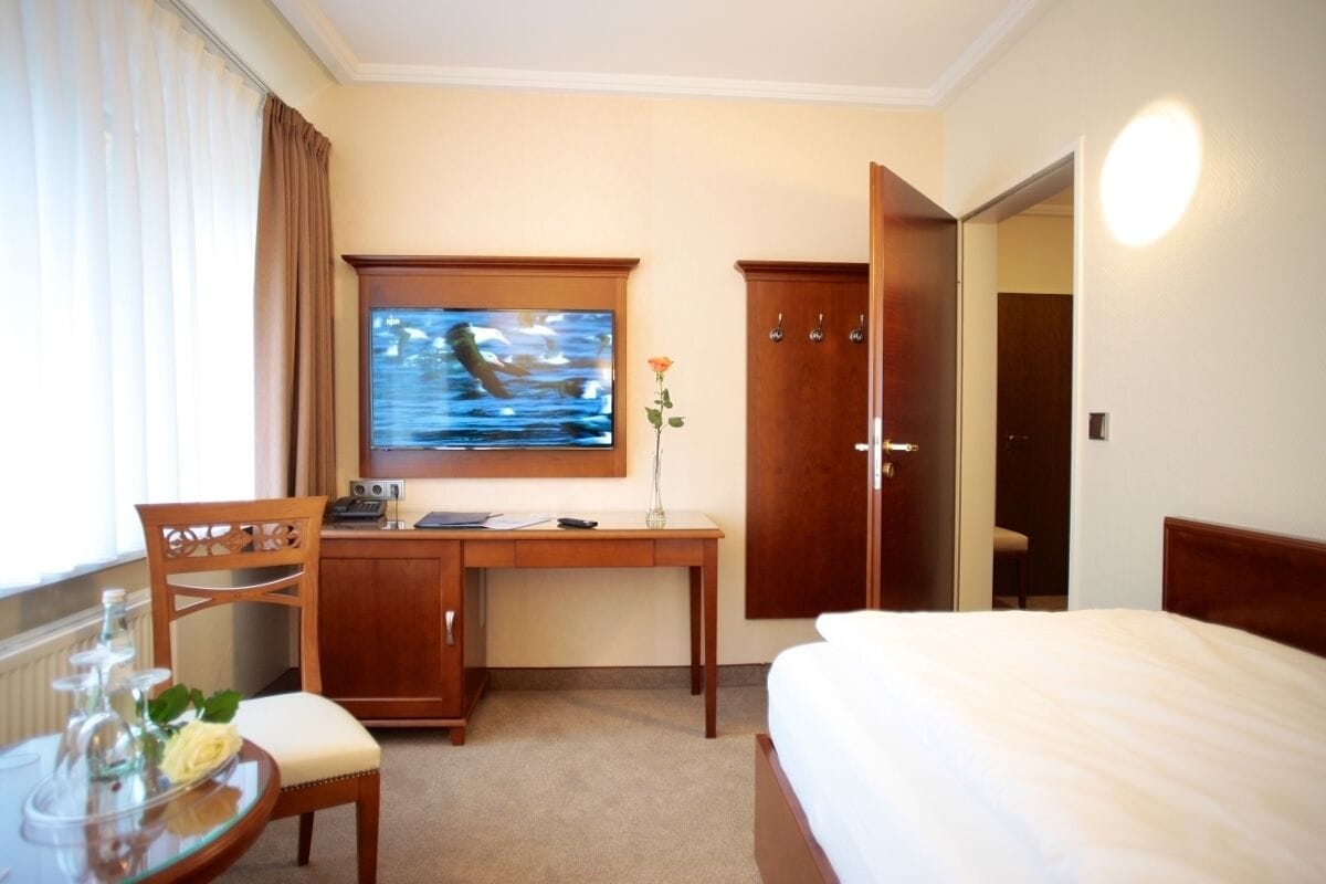 A hotel room with a bed, a desk with a computer, a chair, a TV mounted on the wall, and a door.