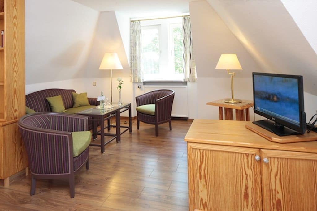 A cozy room with a sofa, two chairs, a small table, a TV on a wooden cabinet, and a window with curtains.