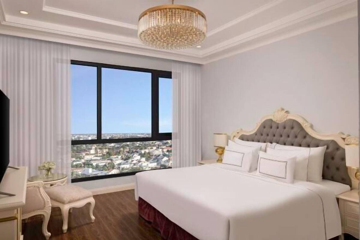Une chambre d’hôtel avec un grand lit, un lustre, une vue sur la ville et une petite table avec une lampe.