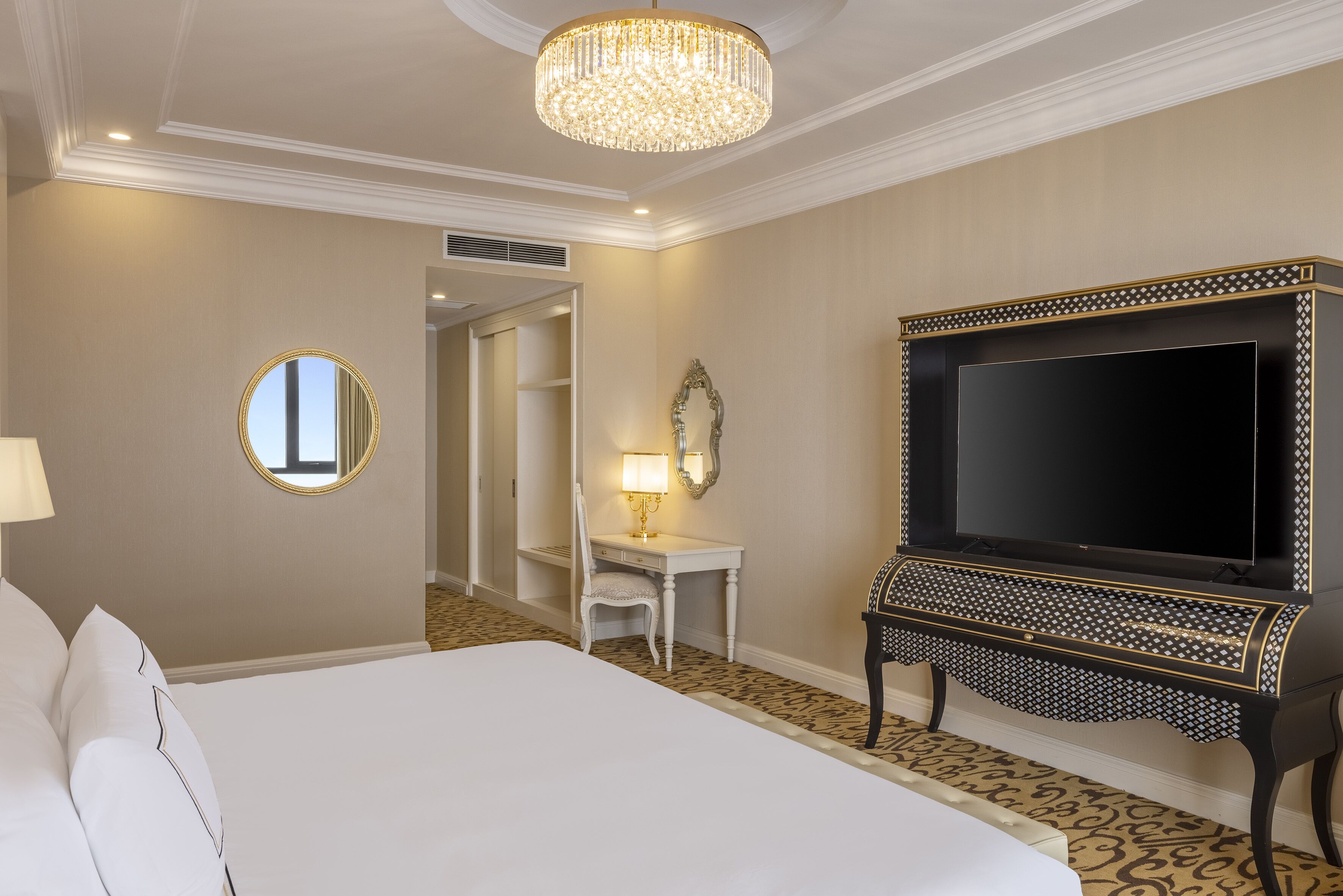 A hotel room with a large bed, a TV on a black stand, a desk with a lamp, and a mirror on the wall.
