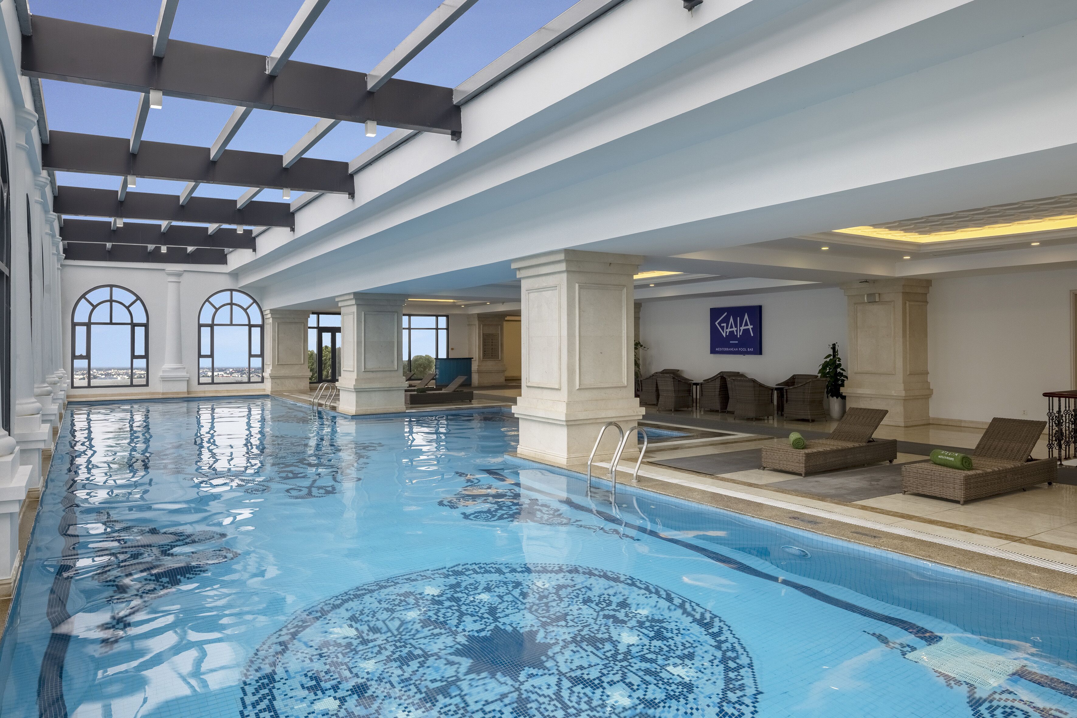 Een groot binnenzwembad met helderblauw water, een plafond met dakramen en een loungehoek met stoelen en een bank.