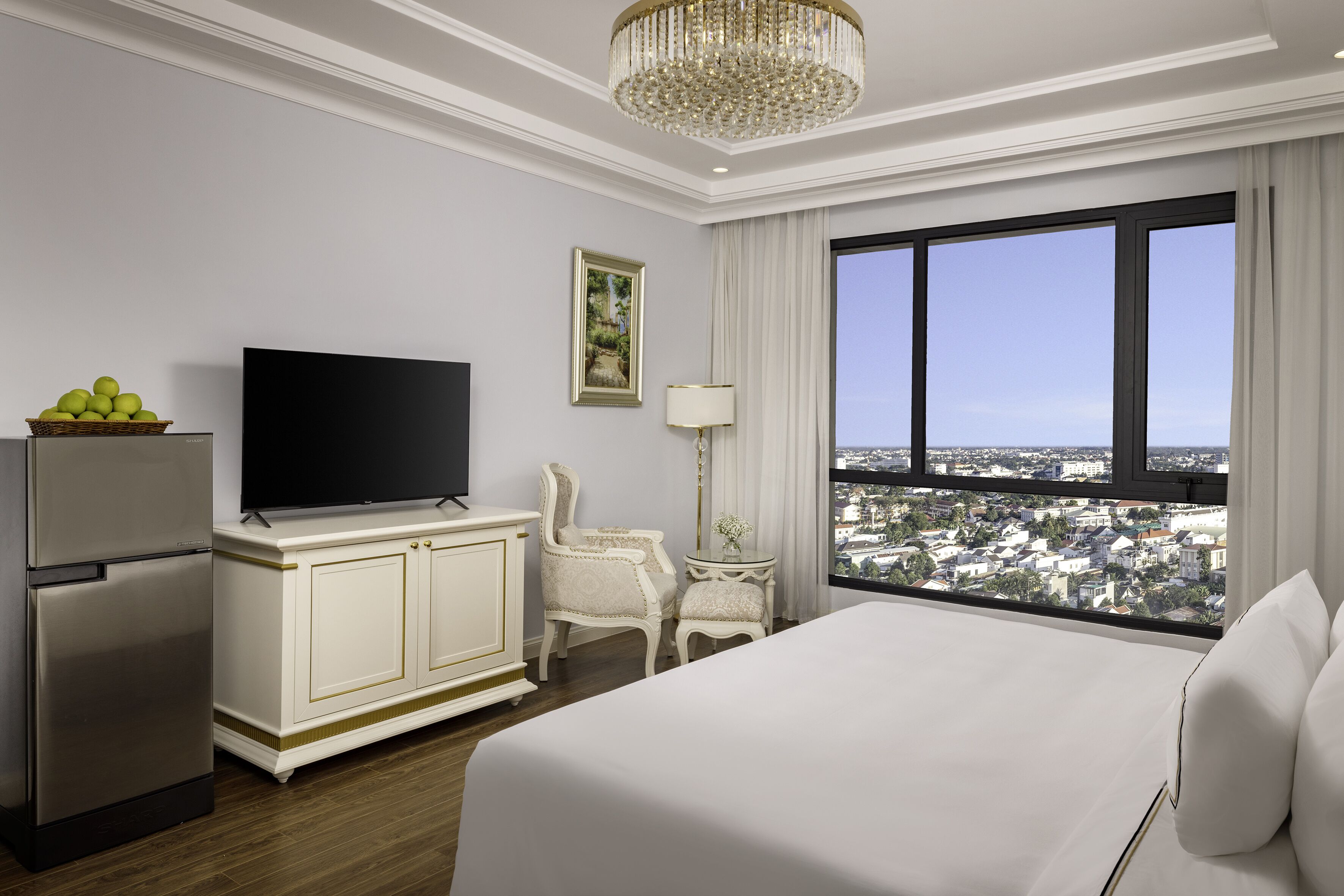Habitación de hotel con una cama grande, televisión, refrigerador, mesa de comedor, silla, lámpara y una ventana amplia con vista a la ciudad.