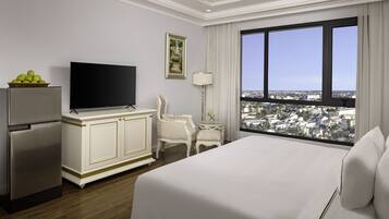 Habitación de hotel con una cama grande, televisión, refrigerador, mesa de comedor, silla, lámpara y una ventana amplia con vista a la ciudad.