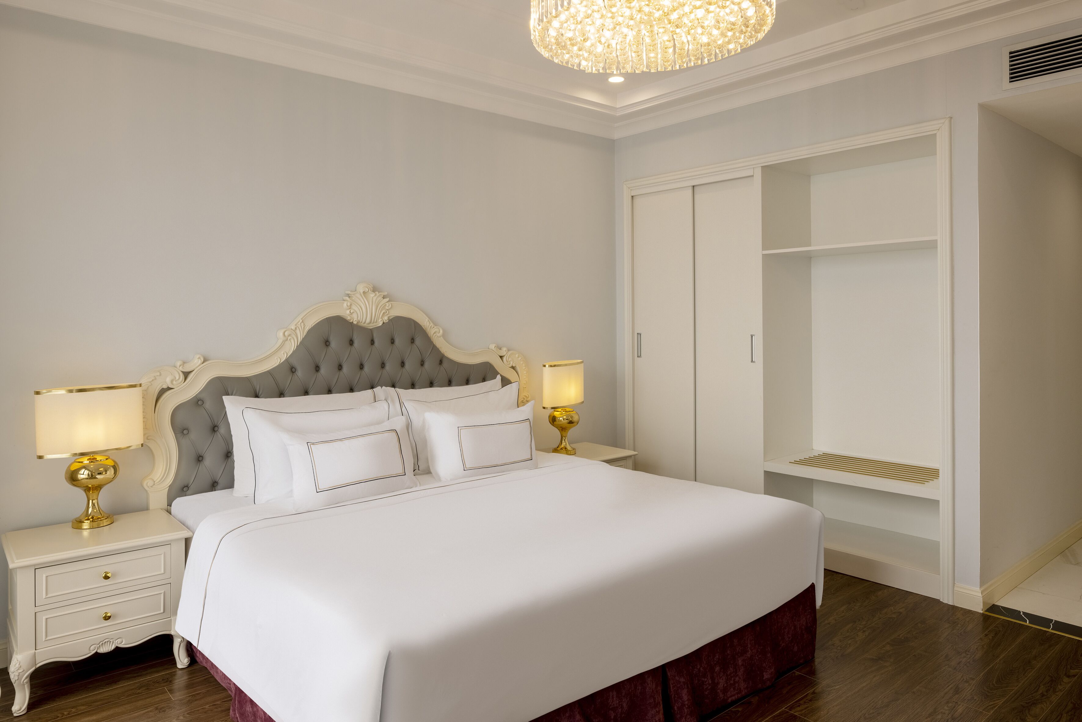 A neatly arranged bedroom with a large bed, two bedside tables with lamps, a wardrobe, and a chandelier.