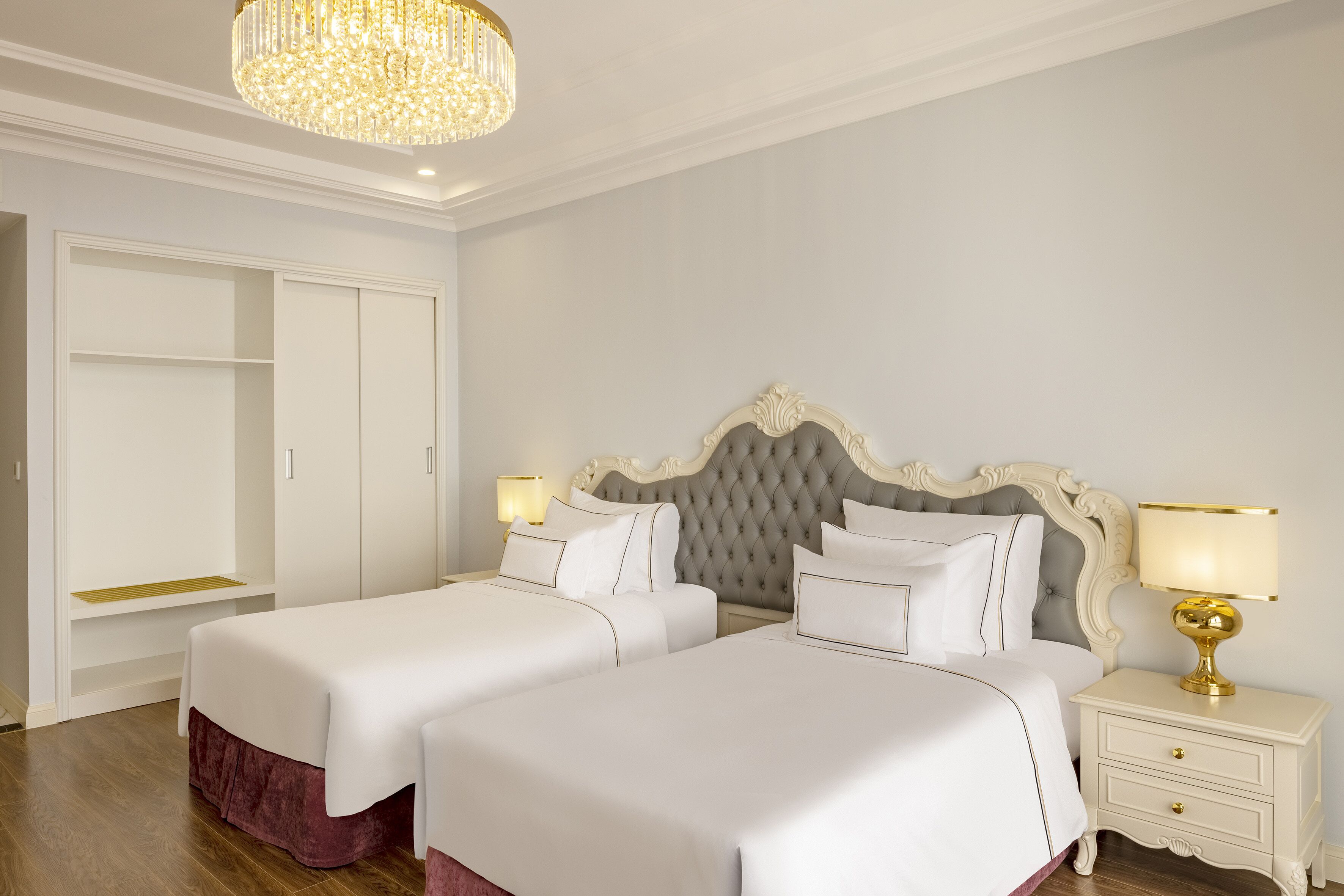Een hotelkamer met twee bedden, een kroonluchter, een kledingkast en een nachtkastje met een lamp.