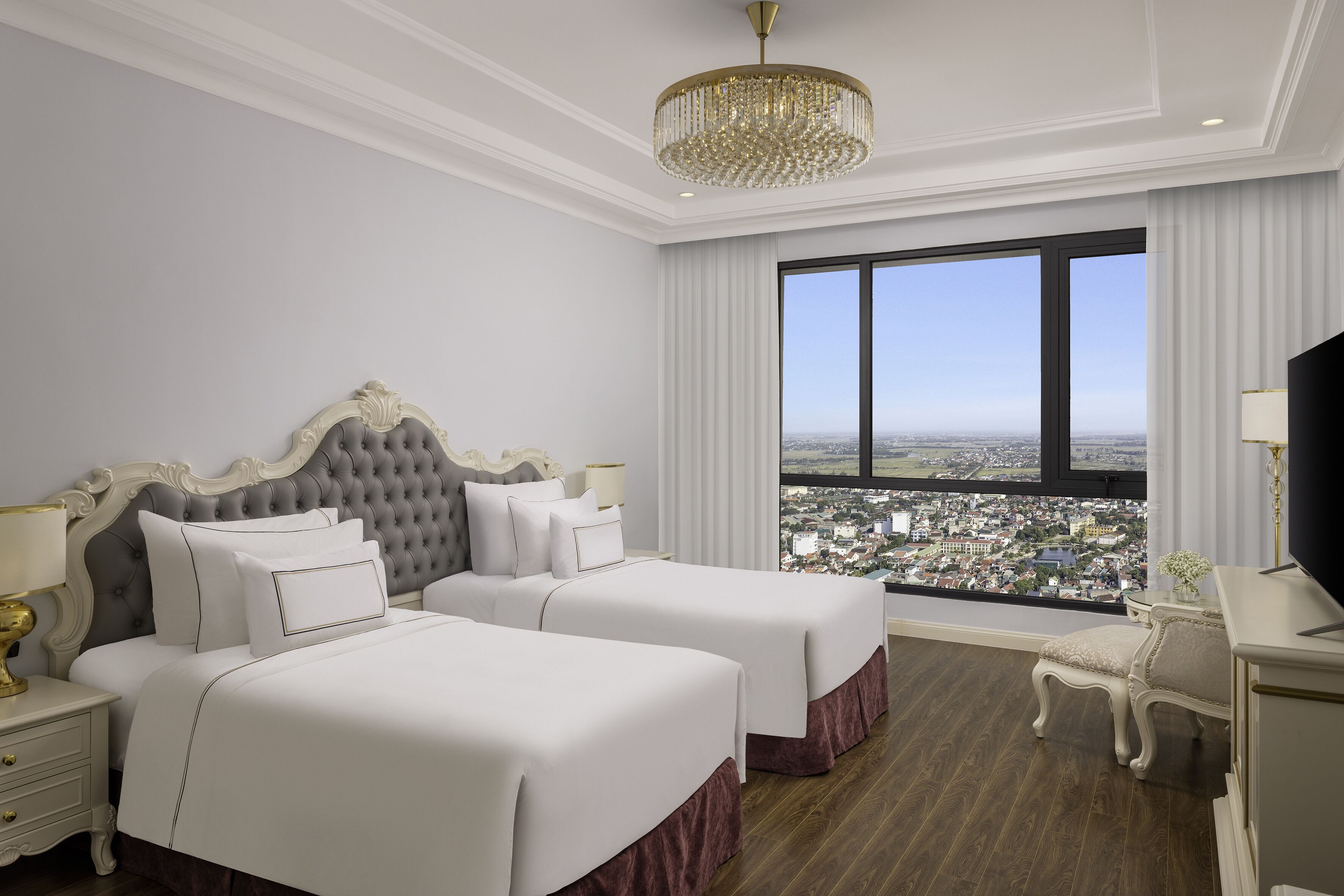 Habitación de hotel con dos camas, un candelabro, un ventanal grande con vista a la ciudad y un escritorio con lámpara.
