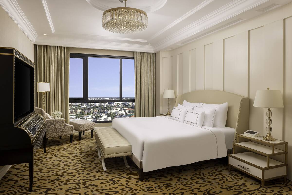Une chambre d’hôtel avec un grand lit, une télévision, un piano, un banc, une table de chevet avec une lampe et une vue sur le paysage urbain par une grande fenêtre.