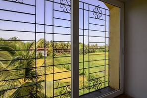 A window with metal bars and a view of a green landscape with palm trees and a building in the distance. - TripThrill Prudential Pristine Apartment (Colva)