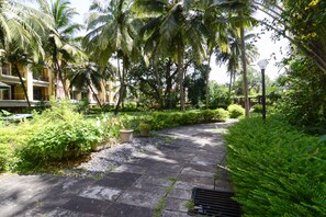 A pathway lined with palm trees and lush greenery, leading to a building with balconies. - TripThrill Prudential Pristine Apartment (Colva)